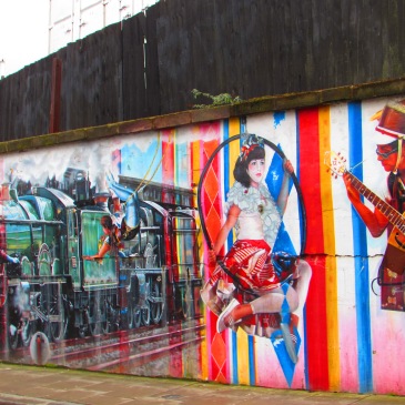 Three graffiti paintings, a green steam train, a pale woman with dark hair and a white top and red sarong swinging on a suspended hoop and a man with a hat playing an acoustic guitar